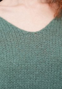 Πλεκτό πουλόβερ σε απαλή πράσινη απόχρωση, με λαιμόκοψη σε σχήμα V. Υφή υφάσματος με ορατές ραφές και ανοιχτό πλέξιμο.