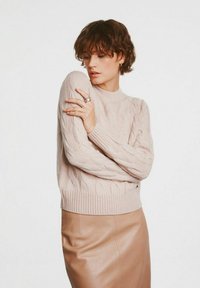 Jasnoróżowy sweter w warkocz z okrągłym dekoltem i ściągaczem, w połączeniu z brązową skórzaną spódnicą. Widoczny jest detal fakturowanej tkaniny.