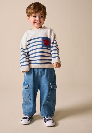 Jeune garçon souriant portant un pull rayé blanc et bleu avec un crabe rouge sur la poche, un jean cargo bleu large et des baskets bleu marine.