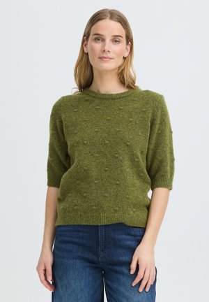 Frau mit hellbraunen Haaren, die einen strukturierten grünen Kurzarmpullover und blaue Jeans trägt, stehend vor einem schlichten hellen Hintergrund.