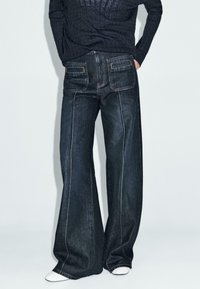 Weite Jeans aus dunklem Denim mit kontrastierenden Nähten, ausgestattet mit zwei Gesäßtaschen und einer mittelhohen Taille. Kombiniert mit weißem Schuhwerk.