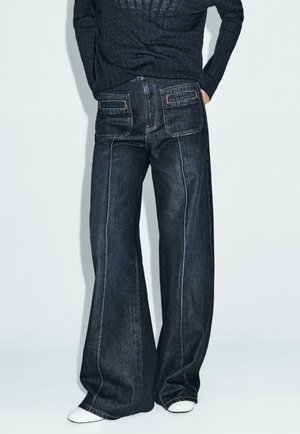 Wijdvallende jeans in donkerdenim met contrastrijk stiksels, voorzien van twee achterzakken en een gemiddelde taillehoogte. Gecombineerd met witte schoenen.