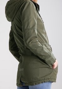 Olivově zelená bunda s kapucí, stahovaným pasem, dlouhými rukávy, předními kapsami a zipovým zapínáním, s hladkým povrchem látky.