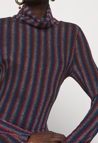 Skinande, kroppsnära höghalspartier med vertikala ränder i rött, blått och metalliska nyanser. Långa ärmar och en mjuk textur.