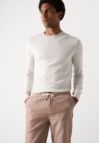Chemise à manches longues crème, col côtelé, petit détail brodé jaune, associée à un pantalon à cordon de serrage marron clair, en tissu texturé.