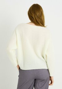 Suéter blanco de punto ligeramente texturizado con un ajuste relajado y hombros caídos, que presenta un escote amplio y un dobladillo acanalado. Combinado con pantalones de rayas grises.