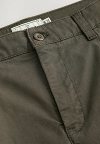 Olivgrüne Baumwollhose mit strukturierter Oberfläche, ausgestattet mit einem Knopfverschluss und einem Etikett im Bund.