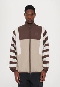 Hnědá a béžová bunda na zip s bílými diagonálními a pruhovanými vzory, s vysokým límcem a elastickými manžetami. Na hrudi logo EA7.