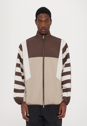 Zip-front jacket featuring brown, beige, and white color blocks with striped sleeves. Made from lightweight material with an elastic hem.