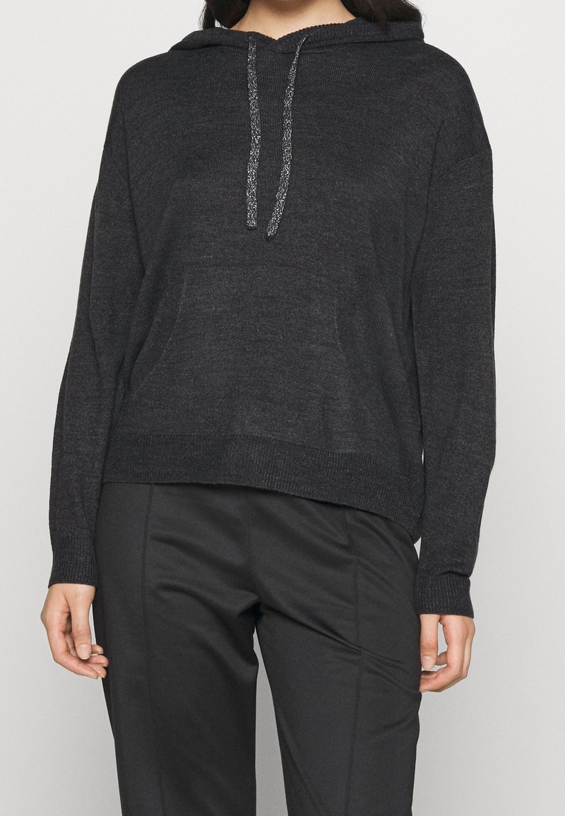Femme portant un sweat à capuche en maille charbon foncé avec poche avant et cordons, associé à un pantalon noir sur mesure sur un fond uni.