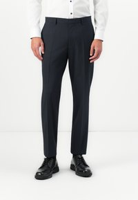 Man wearing black dress pants, black dress shoes, and a white button-up shirt standing against a plain white background.