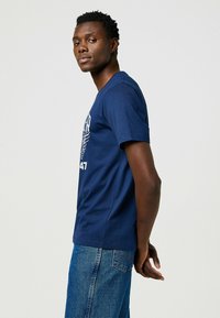 T-shirt blu navy a maniche corte, con scollo rotondo, caratterizzato da una stampa grafica bianca. Indossato con jeans di denim blu chiaro, design semplice.