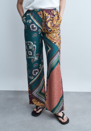 Person wearing multicolored wide-leg pants with paisley and geometric patterns, white top, black flip-flops, hands in pockets, standing on gray floor.