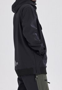 Personne portant une veste noire à capuche, main dans la poche, associée à un pantalon noir avec un panneau de poche zippé vert olive.