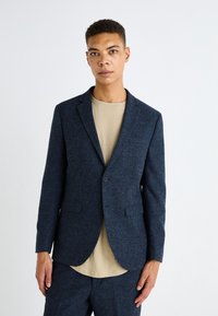 Blazer blu scuro realizzato in tessuto testurizzato, caratterizzato da una chiusura con un bottone singolo, rever a punta e due tasche frontali. Indossato sopra una camicia beige.