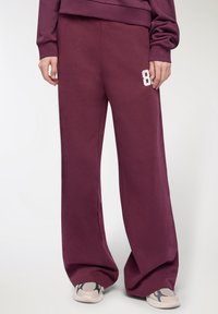 Osoba ubrana w burgundowe szerokie spodnie dresowe z białym nadrukiem "89" na lewym udzie oraz beżowe sneakersy z czarnymi akcentami.