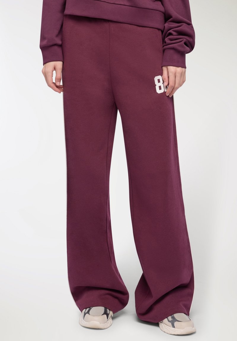 Osoba ubrana w burgundowe szerokie spodnie dresowe z białym nadrukiem "89" na lewym udzie oraz beżowe sneakersy z czarnymi akcentami.