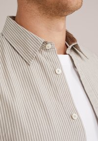 Lichte beige gestreepte shirt met een knoopband en twee zichtbare knopen. De stof heeft een grove textuur met een gladde afwerking.