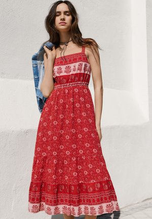 Nainen punaisessa kuvioidussa hihattomassa mekossa pitää sinistä farkkutakkia olkapäänsä päällä ja seisoo valkoista seinää vasten ulkona.