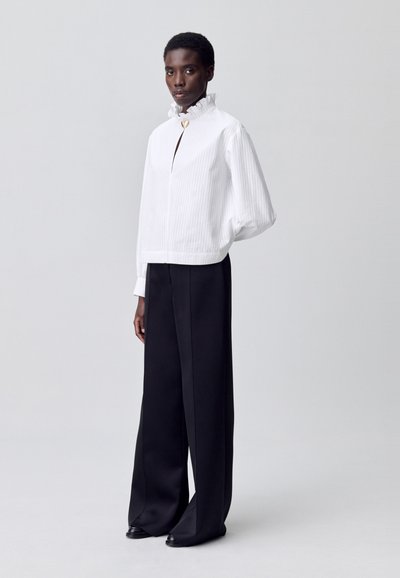 Modelo posa frente a un fondo gris claro, vistiendo una blusa blanca de cuello alto con ribetes y un broche dorado, junto con pantalones anchos negros.