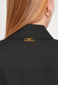 Black shirt with a structured collar, featuring a gold MK logo pin at the back. Smooth texture and simple design with minimal stitching.