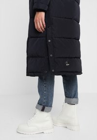 Osoba ubrana w długi czarny pikowany płaszcz, podwinięte niebieskie dżinsy oraz białe sznurowane botki, stojąca na neutralnym tle.