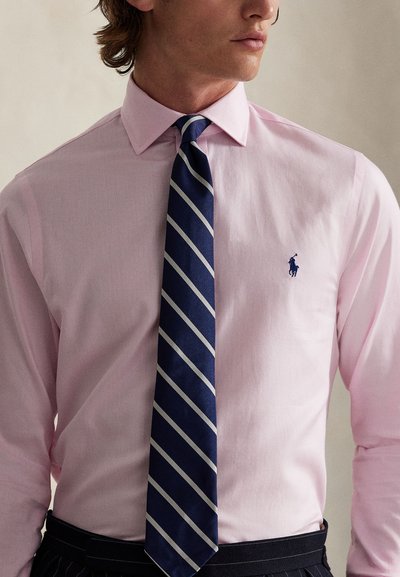 Rožinė marškinėliai su apykakle, kuriuose yra mažas mėlynas logotipas. Derinama su tamsiai mėlyna kaklaraiščiu, dryžuotu baltais ir mėlynais atspalviais.