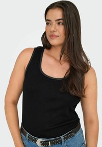 Vrouw met lang bruin haar, gekleed in een zwart zonder mouwen top en blauwe jeans met een zwarte gestifte riem, kijkt opzij tegen een effen achtergrond.