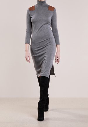 Mannequin portant une robe grise à manches longues et col roulé, avec des patchs marron aux épaules et des bottes noires montant jusqu'aux genoux, marchant vers l'avant.