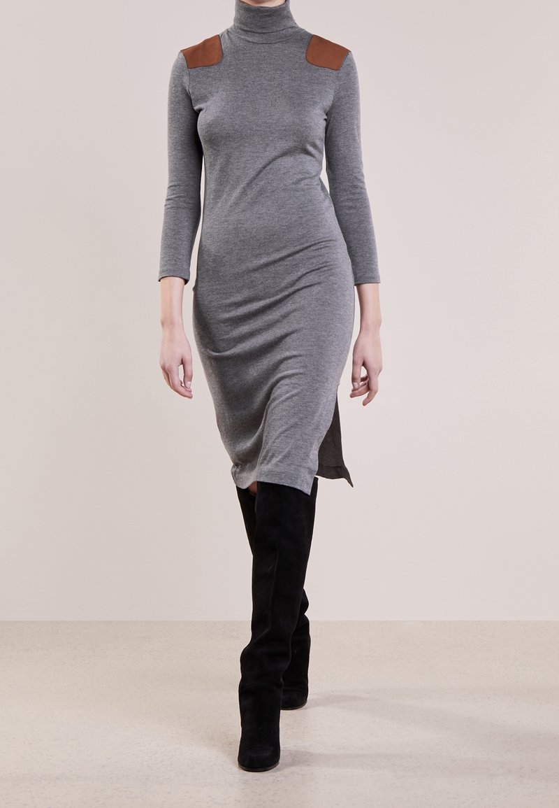 Mannequin portant une robe grise à manches longues et col roulé, avec des patchs marron aux épaules et des bottes noires montant jusqu'aux genoux, marchant vers l'avant.