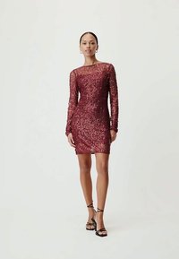 Mulher vestindo um vestido justo de manga longa em lantejoulas burgundy e sandálias de salto alto pretas com tiras, de pé contra um fundo simples.