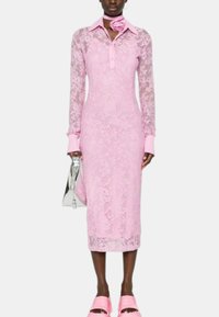 Abito in pizzo rosa con maniche lunghe e colletto, caratterizzato da una texture floreale. Abbinato a una borsa argentata e calzature rosa.