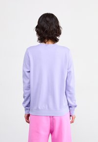 Sweatshirt lilas clair avec des manches longues, des poignets et un ourlet côtelés. Texture lisse, design col rond, associé à un pantalon rose.