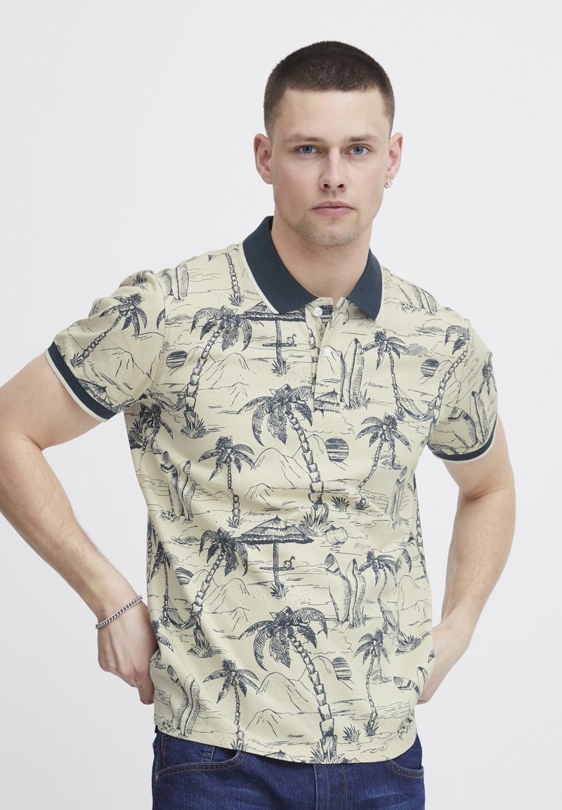Polo tričko s golierom v béžovej farbe, so svetlomodrým lemovaním. S potlačou paliem a tropickej scenérie v tmavomodrej farbe. Krátke rukávy, voľný strih.