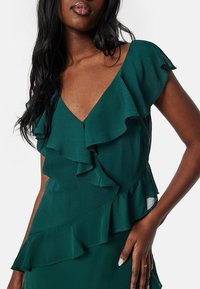 Tyrkysové šaty s volánovými krátkými rukávy a vrstvenými detaily volánů po obvodu dekoltu a pasu, vyrobené z lehké strukturované tkaniny.