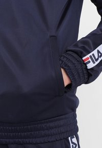 Nahaufnahme einer Person, die einen dunkelblauen FILA-Trainingsanzug trägt, mit der Hand in der Seitentasche und elastischer Manschette am Ärmel.