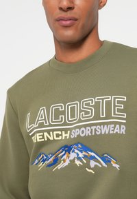 Felpa verde oliva in cotone; presenta una grande grafica di montagna ricamata in blu, arancione e bianco, con testo in grassetto.