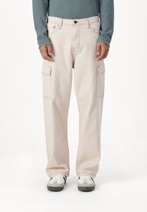 Mężczyzna ubrany w beżowe spodnie cargo z kieszeniami bocznymi, turkusową bluzę z długim rękawem oraz białe trampki, stojący na białym tle.