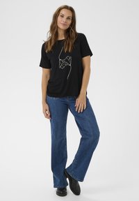 Nő áll fehér háttér előtt, fekete pólót visel, amelyen vonalrajzos virágmintázat található, kék farmert és fekete csizmát visel.