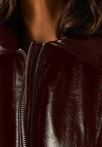 Veste en cuir bordeaux à finition brillante, dotée d'un col montant et d'une fermeture éclair argentée visible sur le devant. Apparence texturée.