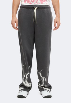 Homme portant un pantalon de survêtement noir avec des motifs d'éclairs blancs près des chevilles, des baskets blanches et un t-shirt blanc avec un texte rose.