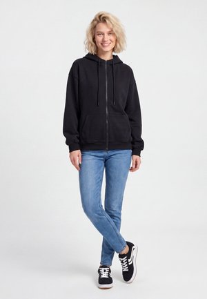 Kvinde med lyst hår iført en sort hættetrøje med lynlås, lyseblå jeans og sorte sneakers, stående mod en ensfarvet lys baggrund.