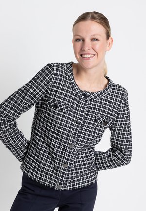 Veste en tweed noire et blanche, avec un col rond, deux poches poitrine avec rabats et des boutons noirs sur le devant.