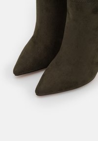 Olivově zelené semišové kotníkové boty s špičatou špičkou a stiletto podpatkem. Hladký povrch s bezšvovým detailem podél horního okraje.