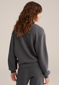 Pelēks pārmērīgs krekls ar nokritušām plecu šuvēm, ribota apakšmala un gluda tekstūra, apvienots ar atbilstošām pelēkām biksēm neitrālā vidē.