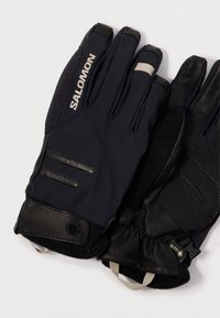 Melnas cimdi, kas izgatavoti no auduma un ādas maisījuma, ar teksturētu satvērienu, elastīgu aproci un logo detaļu augšpusē. Akcenti pelēkā krāsā.