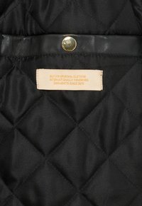 Black quilted fabric with a glossy finish, featuring a gold snap button and a woven label stating "BLK/VIS ORIGINAL CLOTHING."
