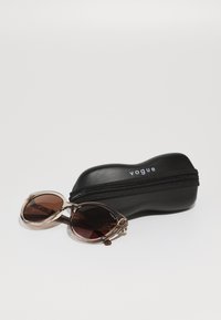 Ochelari de soare cu rame transparente de culoare maronie și lentile maro închis, însoțiți de o husă neagră cu fermoar, etichetată "vogue."
