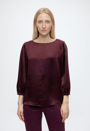 Mujer con el pelo rubio liso lleva una blusa burdeos brillante de manga tres cuartos y pantalones burdeos a juego, de pie frente a un fondo liso.