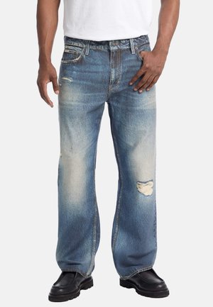 Vaqueros de denim azul claro con un ajuste relajado, que presentan parches desgastados y un acabado deslavado. Se combinan con zapatos negros y una camiseta blanca.